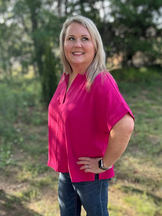 Woman wearing a pink shirt and blue jeans standing outdoors with trees in the background