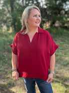 Woman wearing a red shirt and blue jeans standing outdoors with trees in the background
