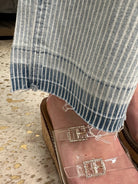 Close-up of striped jeans and clear sandals with rhinestone buckles on a textured floor.