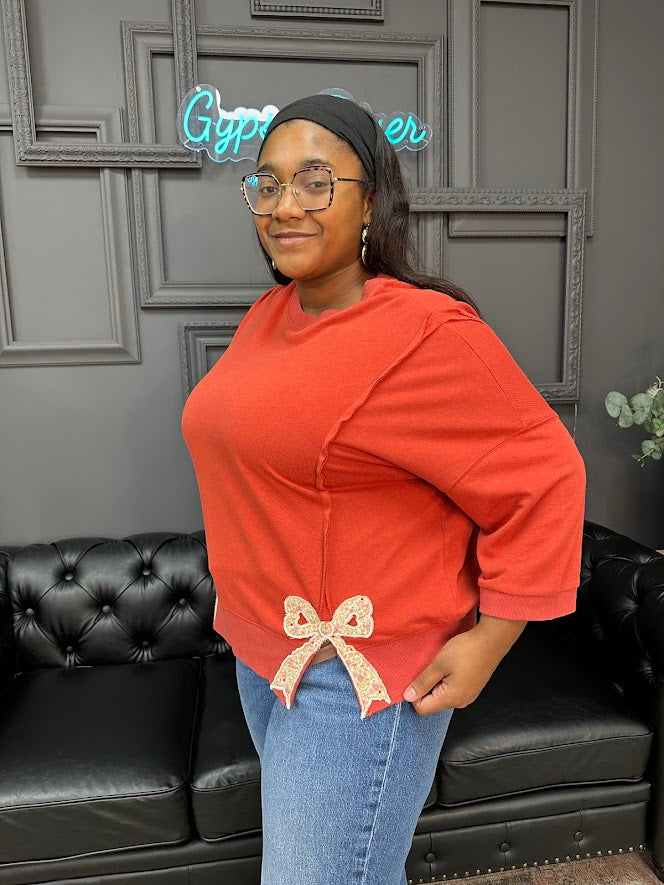 Woman wearing an orange sweater with a bow detail, standing in front of a black leather couch and gray wall with neon sign.
