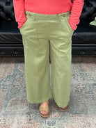 Woman in a pink sweater and green pants standing in a room with a black couch and decorative items.