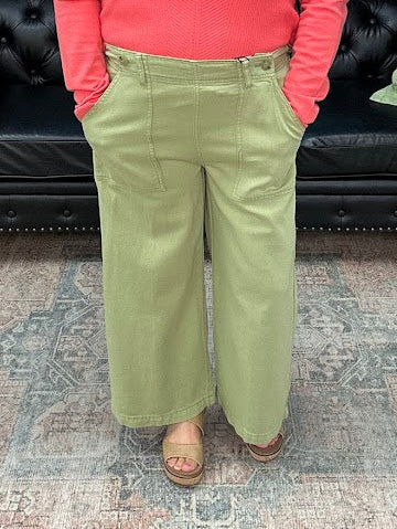 Woman in a pink sweater and green pants standing in a room with a black couch and decorative items.