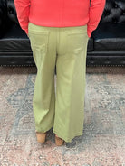 Person wearing a coral sweater and green pants standing in a room with a black leather couch and floral decorations.