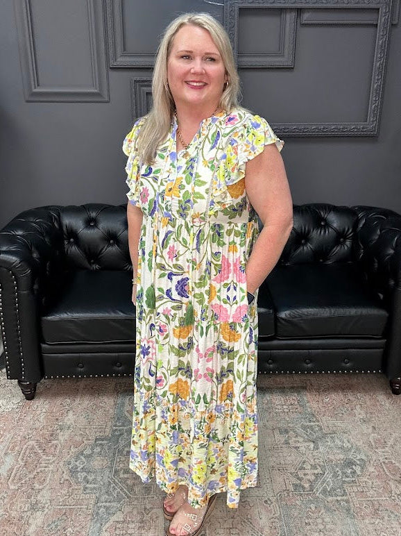 Woman in a floral dress standing in front of black leather chairs with a dark gray wall and neon sign in the background.