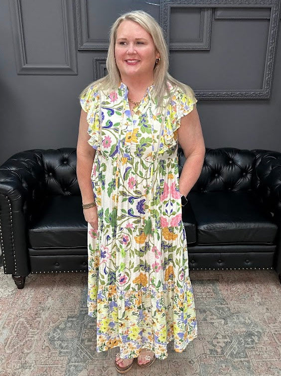 Woman in a floral dress standing in front of a black leather couch with a gray wall and neon sign in the background.