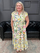 Woman in a floral dress standing in front of a black leather couch with a gray wall and neon sign in the background.