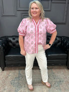Woman wearing a pink floral blouse and white pants standing in a room with a black leather couch.