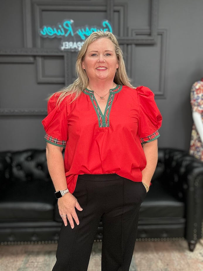 Woman in a red blouse with green accents standing in a room with a dark gray wall and black leather couch.