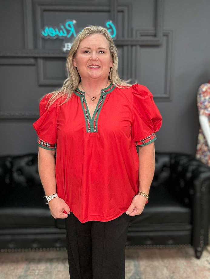 Woman wearing a red blouse with green accents in a room with a black leather couch and decorative wall.