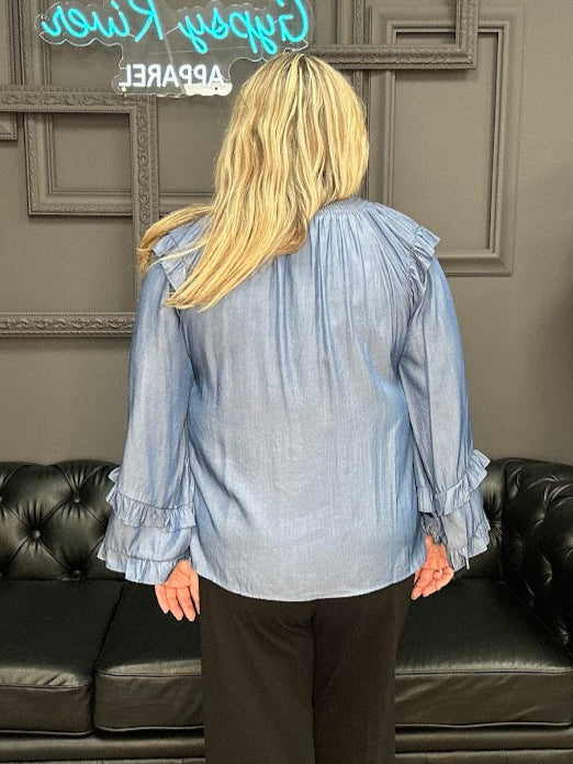 Person wearing a light blue blouse with ruffled sleeves in a room with a black leather couch and decorative wall.