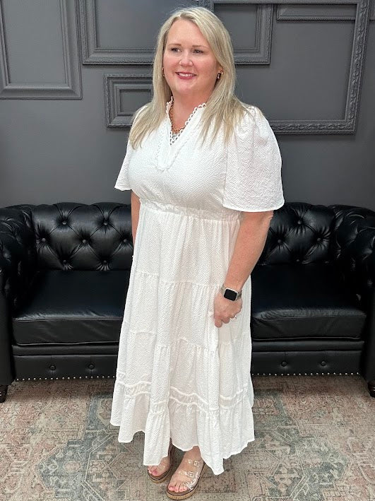 Woman in a white dress standing in front of a black leather couch with a gray wall and neon sign in the background.