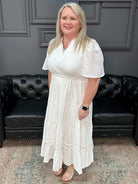 Woman in a white dress standing in front of a black leather couch with a gray wall and neon sign in the background.