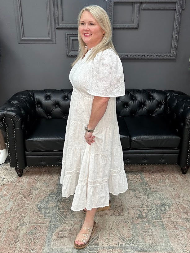 Woman in a white dress standing in a room with black leather couches and gray walls.