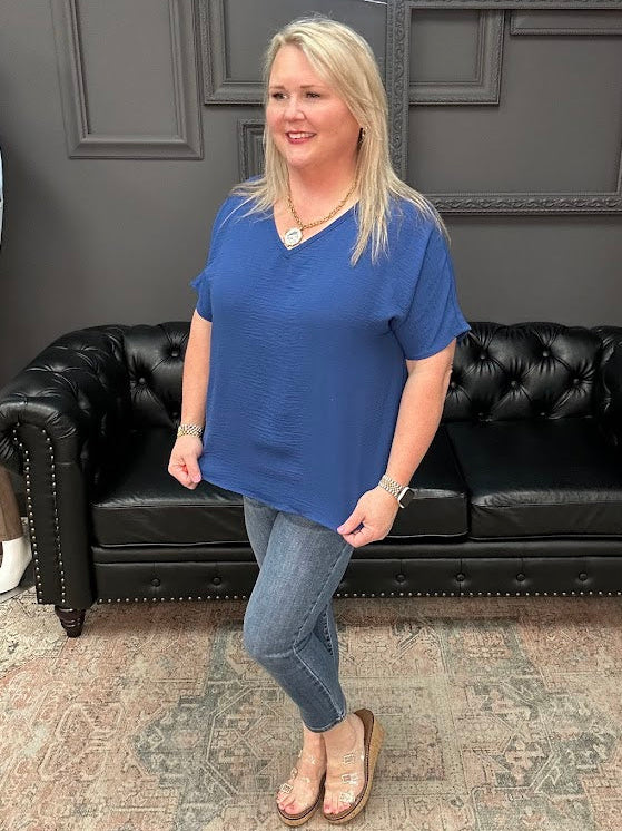 Woman in a blue shirt standing in front of a black leather couch with a gray wall and neon sign in the background.
