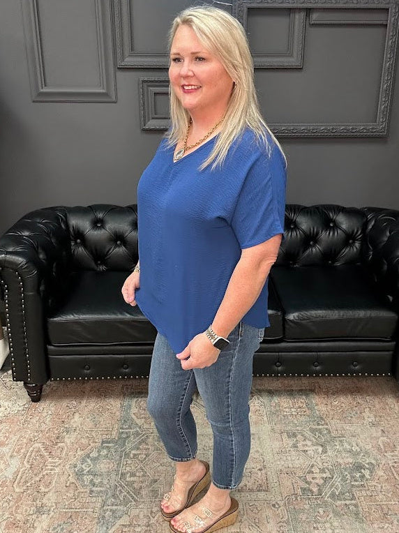 Woman in a blue shirt standing in front of a black leather couch with a gray wall and neon sign in the background.