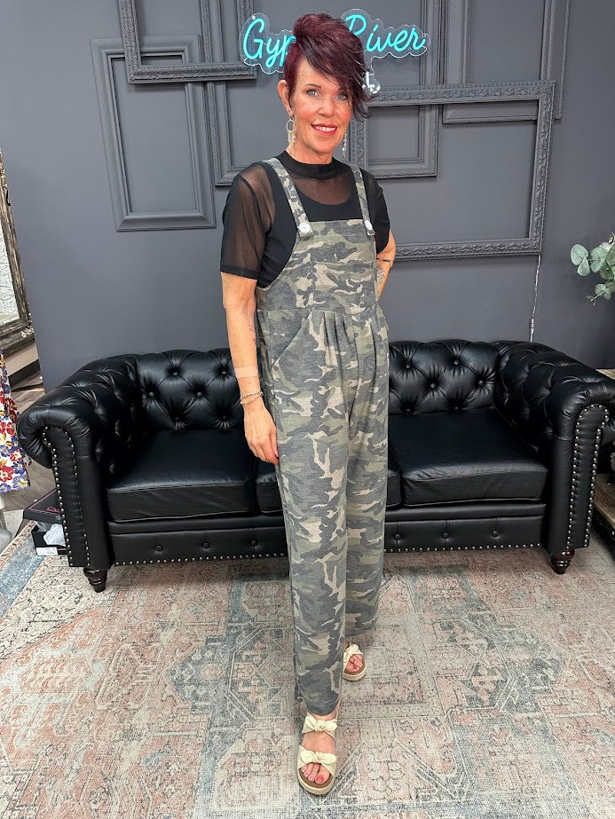 Woman wearing camouflage overalls standing in a room with a black sofa and gray walls.