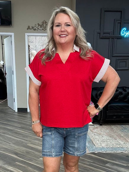 Woman in a red shirt and denim shorts standing in a store with a television and neon sign in the background.